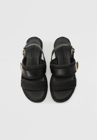 Sorte sandaler med to vævede stropper, et krydset design og en guldtonet spændeaccent. Flad sål og teksturerede kanter.