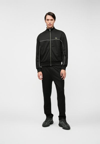 Survêtements noirs comprenant une veste zippée avec des pipings blancs et un pantalon assorti. Tissu texturé avec une coupe ajustée et un accent de logo.