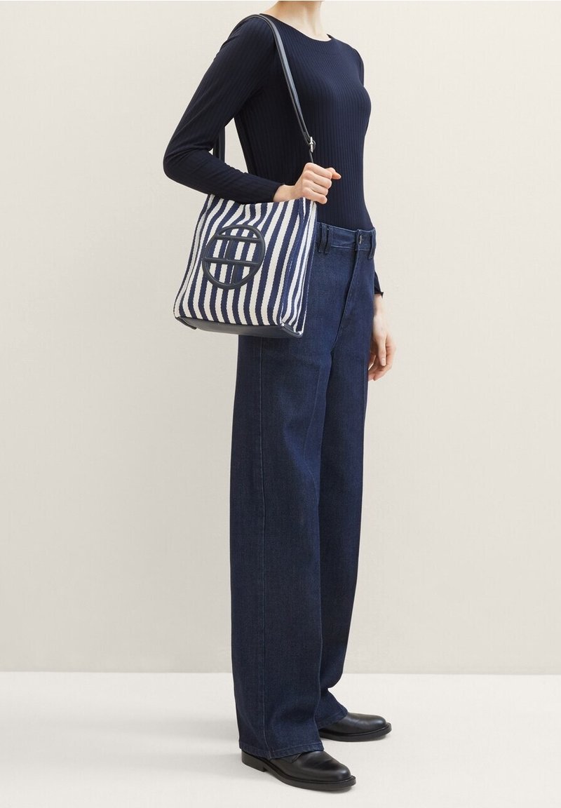 Borsa a spalla in canvas a righe con righe verticali blu scuro e bianche, dettaglio rotondo con logo, tracolla nera; abbinata a pantaloni larghi blu navy.