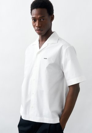 Άνδρας που φοράει κοντομάνικο λευκό πουκάμισο με κουμπιά και λογότυπο "HUGO", με τα χέρια στις τσέπες, μπροστά σε ένα απλό φωτεινό φόντο.