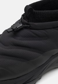 Scarpa nera isolata con superficie texturizzata, design aderente, polsino elastico alla caviglia e un toggle in plastica per una regolazione sicura.