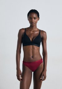 Zwarte bralette met een V-halsontwerp en dunne bandjes, gecombineerd met rode panties met een zachte tailleband en logodetail. Strakke pasvorm.