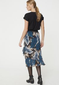 Schwarzes Kurzarmoberteil kombiniert mit einem blau-beigen Rock mit Federdesign, der Rüschendetails und einen schwarzen Bund hat. Sie trägt schwarze Stiefeletten.