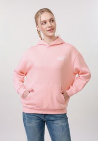 Jeune femme portant un sweat à capuche rose avec poches et un jean bleu, regardant sur le côté avec les mains dans les poches du sweat.
