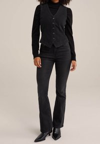 Černá denimová vesta s výstřihem do V a pěti knoflíky, v kombinaci s černým topem s dlouhým rukávem a rozšířenými černými džíny. Hladká textura, přiléhavý střih.