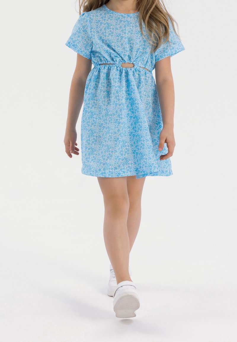 Jeune fille marchant dans une robe courte bleue à fleurs avec une découpe devant, portant des baskets blanches sur fond blanc uni.