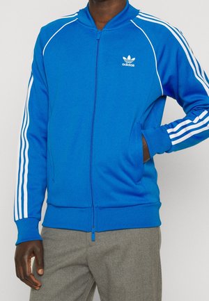 Mężczyzna w niebieskiej kurtce sportowej Adidas z białymi paskami na rękawach i szarych spodniach, stojący z jedną ręką w kieszeni.