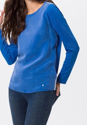Vrouw met een blauw shirt met lange mouwen en donkerblauwe spijkerbroek, hand die haar haar aanraakt, tegen een effen lichte achtergrond.