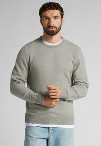 Jasnoszary sweter z dzianiny z okrągłym dekoltem, prążkowanymi mankietami i dolnym wykończeniem, w zestawieniu z białą podkoszulką. Tekstura wydaje się miękka i gładka.