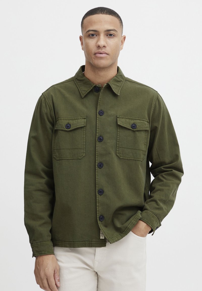 Olive green utility jacket in cotton fabric, featuring button closure, two chest pockets, and a structured collar.