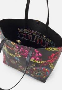 Black tote bag with colorful baroque patterns and "Versace Jeans Couture" text inside, featuring two black handles and gold accents.