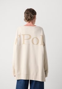 Crème oversized sweatshirt met beige logo print op de rug, ronde halslijn en geribde boorden, gecombineerd met blauwe wijd-gesneden jeans.