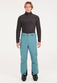 Pánsky lyžiarsky outfit pozostávajúci z čierneho, dlhého rukávu, s výťahom na zips a vodoodpudivých tehlovo modrých nohavíc. Nohavice majú rovný strih a topánky sú čierne.
