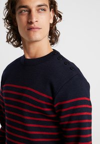 Granatowy sweter z czerwonymi poziomymi paskami, żebrowany dekolt i cztery czarne guziki na lewym ramieniu. Miękka tekstura, swobodny styl.