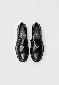 Pantofi loafer din piele lac neagră, cu elemente hardware aurii în formă de logo GUESS pe curea, expuși unul lângă altul pe un fundal alb.