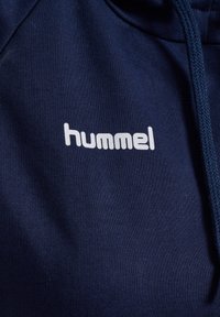 Granatowy materiał na bluzę z kapturem z białym logo „hummel” nadrukowanym na piersi oraz widocznymi granatowymi sznurkami.