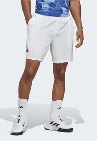Hvide sportsshorts med elastisk talje, sidelommer og sort Adidas-logo. Bæres med hvide sokker med sorte striber og hvide sneakers.
