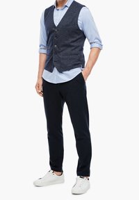 Marineblaues, strukturiertes Weste über einem hellblauen Hemd, kombiniert mit dunklen Hosen und weißen Sneakers. Hat Knöpfe, Taschen und hochgekrempelte Ärmel.