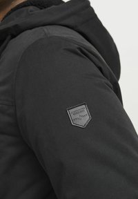 Schwarze Jacke mit strukturierter Oberfläche. Verfügt über ein dreieckiges Logo-Patch auf der Schulter. Das Design umfasst eine Kapuze und Raglanärmel.