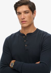 Camisa henley de manga larga en color navy con cuello redondo, cuatro botones marrones y un diseño ajustado. Se lleva con un reloj dorado en la muñeca.