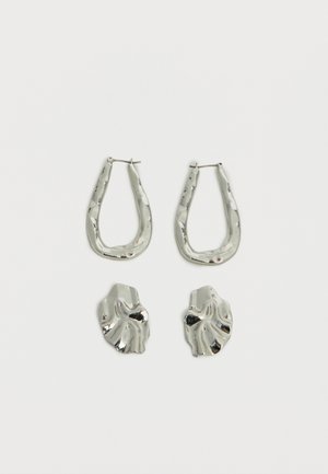 Două perechi de cercei argintii, una cu cercuri ovale neregulate și una cu cercei abstracte, ondulați, pe un fundal alb simplu.