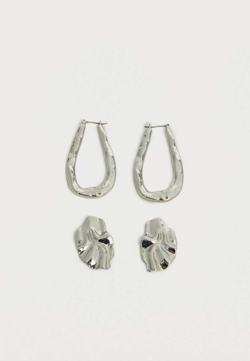 Δύο ζευγάρια σκουλαρίκια σε ασημί τόνο, το ένα με ακανόνιστα οβάλ κρίκους και το άλλο με αφηρημένα, κυματιστά καρφωτά, σε απλό λευκό φόντο.