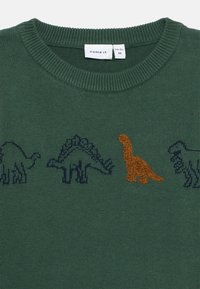 Žalias megztinis su dinozaurų raštais tamsiai mėlyna spalva ir tekstūruotu oranžiniu dinozauru. Klasikinis apvalus kaklas su nertais kraštais.