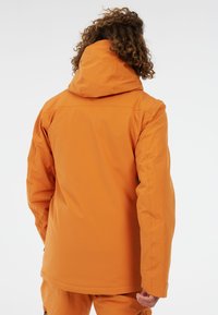 Chaqueta impermeable naranja con capucha, que presenta una textura suave, un ajuste ligeramente holgado y puños ajustables. La vista trasera no muestra logotipos.