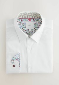 Bela bombažna srajca z teksturirano površino. Notranji ovratnik in manšeta imata cvetlični vzorec v različnih barvah, na manšeti je rdeč gumb.