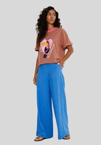 Hnědé tričko s krátkým rukávem a barevným motivem tukana, s růžovým výstřihem, zkombinované s modrými širokými kalhotami a sandály.