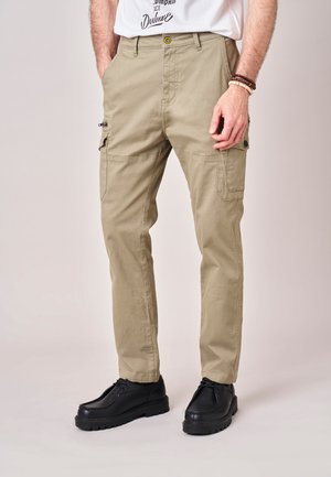 Man draagt beige cargobroek met zijzakken en zwarte veterschoenen, staand tegen een effen achtergrond.