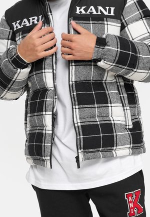 Mains ajustant une doudoune à carreaux noir et blanc avec des écussons logo blancs "KARL KANI" sur la poitrine, portée sur une chemise blanche.