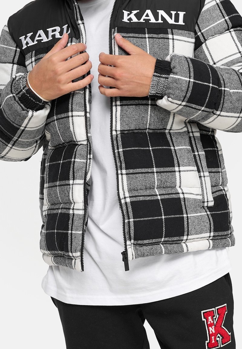 Mains ajustant une doudoune à carreaux noir et blanc avec des écussons logo blancs "KARL KANI" sur la poitrine, portée sur une chemise blanche.