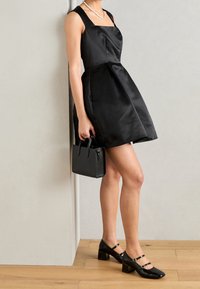 Abito in satin nero con scollatura quadrata e gonna plissettata, abbinato a scarpe con tacco a blocco in pelle lucida nera e una borsa nera strutturata.