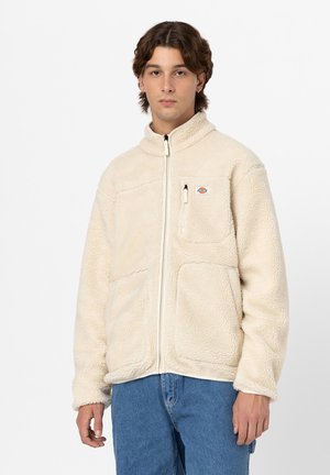 Veste en polaire crème avec un col haut et une fermeture éclair à l'avant, dotée de deux poches latérales et d'une poche poitrine. Tissu doux et texturé.