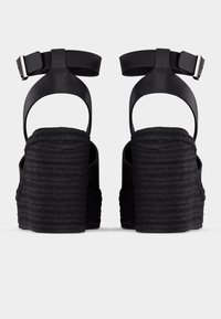 Paire de sandales compensées noires avec des talons tressés épais et des sangles à la cheville avec boucles argentées, vues de dos.