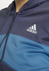 Personne portant un sweat à capuche zippé Adidas bleu marine et bleu avec un logo blanc Adidas visible sur la poitrine.