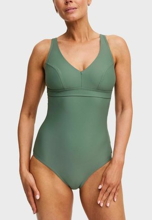 Vrouw draagt een mouwloze, olijfgroene badpak met V-hals, staand met ontspannen armen tegen een effen lichtgrijze achtergrond.