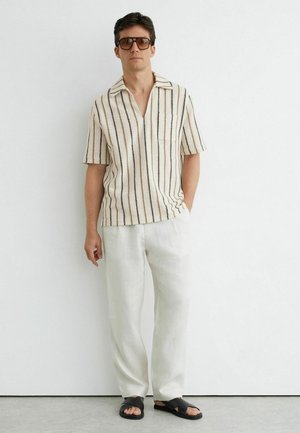 Mann mit gestreiftem kurzärmligem Hemd, weißen Hosen, dunkler Sonnenbrille und schwarzen offenen Sandalen steht vor einer einfachen weißen Wand.