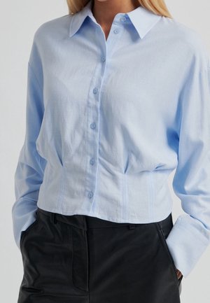 Femme portant une chemise bleu clair à manches longues boutonnée avec col, rentrée dans un pantalon noir taille haute, les mains dans les poches.