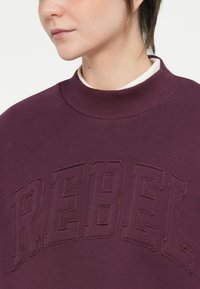 Burgundijas sporta krekls ar ribotu apkakli un izceltiem "REBEL" burtiem. Audumam ir maiga un gluda tekstūra.