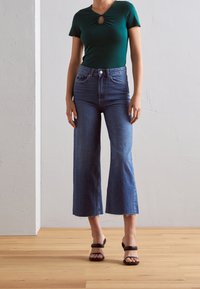 Vihreä lyhythihainen paita, jossa on pisaranmuotoinen avanne, yhdistettynä sinisiin leveälahkeisiin farkkuihin, joissa on rispaantunut helma, ja mustiin korkokenkiin.