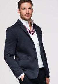 Homme portant un blazer bleu marine sur un pull blanc à col en V avec des rayures bordeaux et bleu marine, posant avec une main dans la poche devant un fond uni.