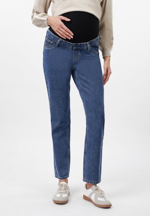 Personne enceinte portant un jean de maternité bleu et des baskets blanches, main posée sur le ventre, haut beige à manches longues visible au-dessus du jean.