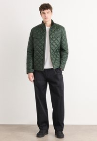 Veste matelassée verte avec col montant, deux poches poitrine et fermeture éclair. Portée sur un t-shirt blanc et un pantalon noir, avec des chaussures noires.