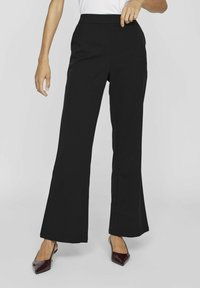 Črne široke hlače z gladko teksturo, visokim pasom in sprednjimi šivi. Počivajo na burgundskih čevljih s koničastim nosom.