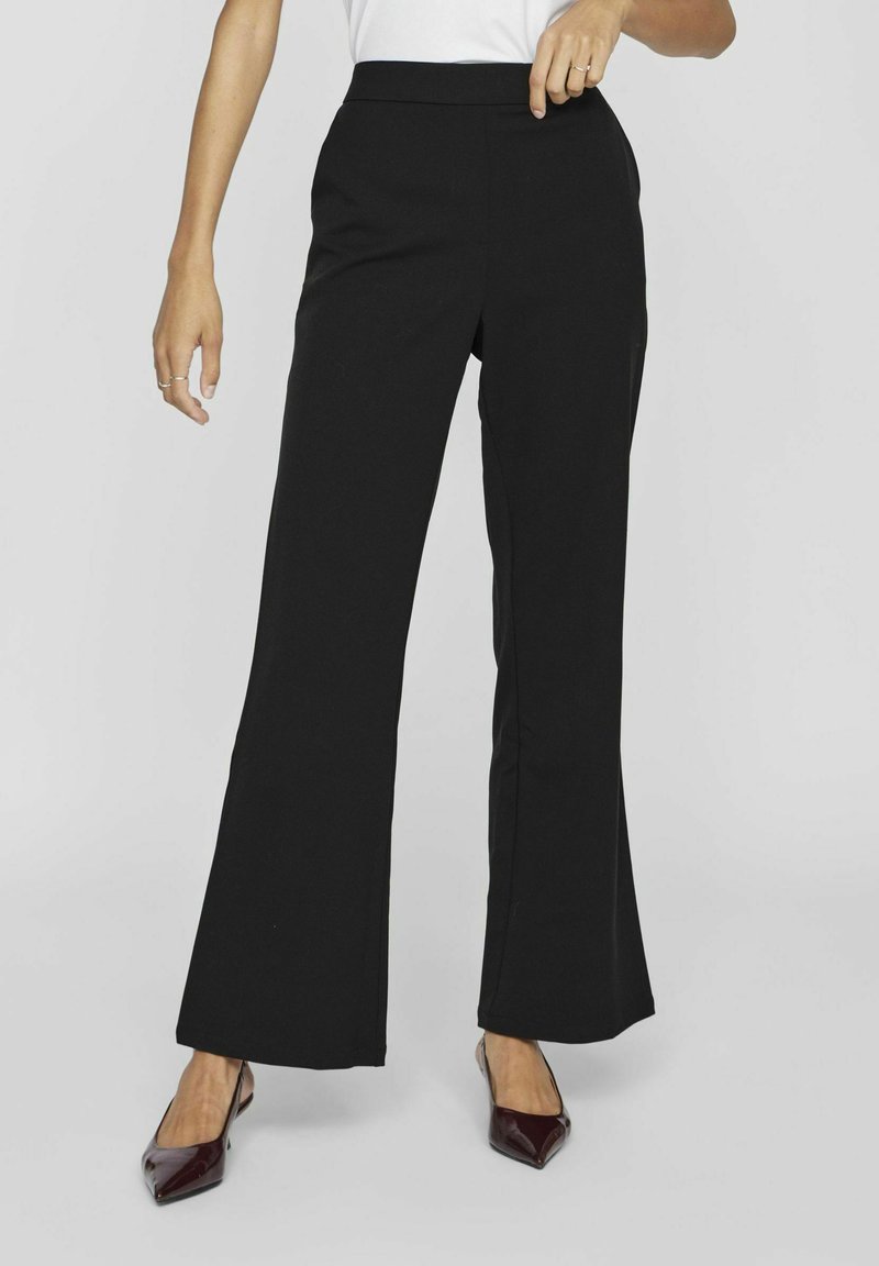 Črne široke hlače z gladko teksturo, visokim pasom in sprednjimi šivi. Počivajo na burgundskih čevljih s koničastim nosom.