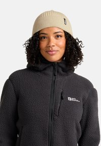 Donna che indossa un berretto a maglia beige e una giacca nera con zip e cappuccio, con il logo Jack Wolfskin sul petto.
