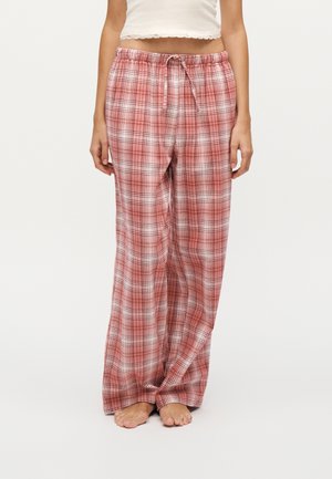 Vrouw draagt losse, rood-wit geruite pyjamabroek met elastische tailleband en trekkoord, staat blootsvoets tegen een witte achtergrond.