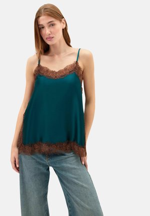 Ung kvinde med glat lysbrunt hår iført en teal satin camisole med brun blondekant og løse blå jeans mod en ensfarvet baggrund.
